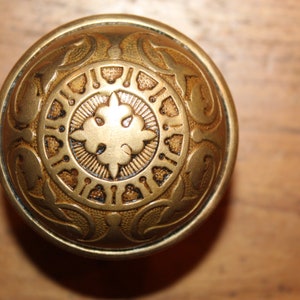 May include: A gold-toned door knob with an intricate, ornate design. The knob has a circular shape and features a central floral motif surrounded by a ring of smaller decorative elements.