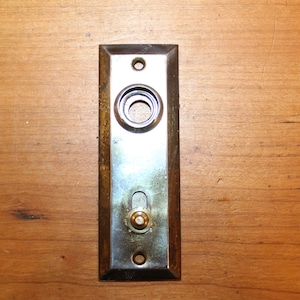 May include: A vintage door viewer with a brass trim and a round peephole. The viewer is mounted on a wooden surface.