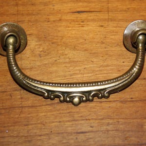 May include: A vintage brass drawer pull with an ornate design. The pull has a curved shape with a decorative center and two round knobs on either end.