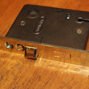 May include: A vintage door lock with a brass-colored faceplate and a dark metal body. The lock features a rectangular design with screw holes and a keyhole. The faceplate has a rectangular strike plate and a protruding latch.