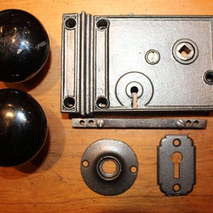 May include: An antique door hardware set, featuring two black round door knobs, a rectangular lock plate, a small round plate, a keyhole cover, and a connecting bar. The set is made of metal and is likely from the early 20th century.