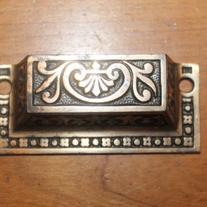 May include: Antique bronze-toned drawer pull with ornate floral and scrollwork detailing. The rectangular handle features a raised central panel with symmetrical designs and is flanked by two circular holes for mounting. A decorative border surrounds the handle.