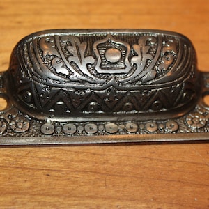 May include: A silver-toned metal drawer pull with an ornate design. The pull has a rectangular base with a curved top and intricate floral and geometric patterns.