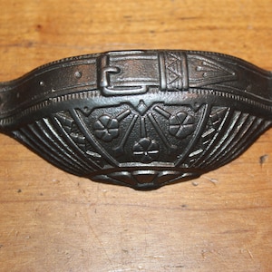 May include: A black cast iron drawer pull with an ornate design featuring a belt buckle and floral motifs.