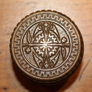 May include: A detailed, circular bronze-colored object with an ornate, symmetrical design. The central motif features a cross-like shape surrounded by floral and geometric patterns. The outer edge has a repeating pattern. The object is likely a decorative element or hardware.