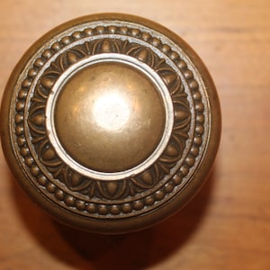 May include: A brass door knob with a decorative, ornate design. The knob has a circular shape with a raised center and a beaded border.
