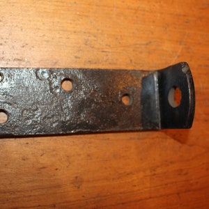 May include: A black metal bracket with four holes and a bent section. The bracket is rusty and has a rough texture.