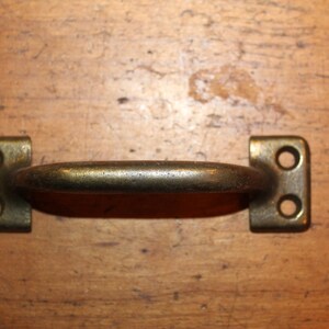 May include: A brass door handle with a rounded shape and two holes on each side. The handle is mounted on a wooden surface.