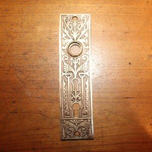 May include: Ornate, rectangular antique door plate with a keyhole and a circular knob opening. The plate is made of a metallic material with an intricate floral and geometric design. The plate is a light bronze color.