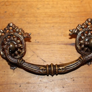 May include: A vintage brass drawer pull with a decorative floral design. The pull has a curved shape and is attached to a wooden surface.