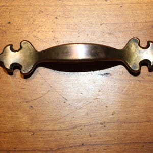 May include: A bronze-colored cabinet pull with decorative ends. The handle is curved and has a smooth, polished finish. The ends feature a stylized, ornate design. The hardware is set against a wooden surface.