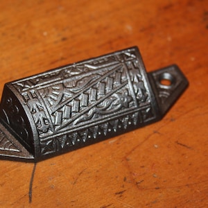May include: A black cast iron drawer pull with an ornate design. The pull has a rectangular shape with rounded edges and a decorative pattern on the top.