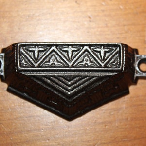 May include: A black cast iron drawer pull with a geometric design. The pull has a rectangular shape with a pointed bottom and two round holes on the sides.