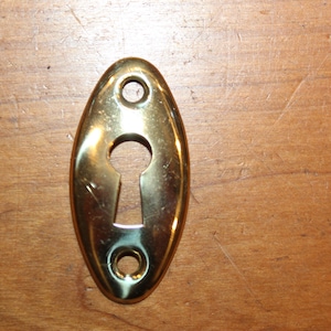 May include: A brass keyhole cover with an oval shape and two small holes on either side.