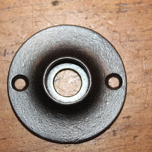 May include: A round, dark-colored metal flange with a central hole and two smaller holes. The flange has a textured surface and is likely used for mounting or connecting components. The object is set against a wooden background.