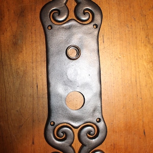 May include: A dark gray, cast iron door plate with decorative scrollwork at the top and bottom. The plate has a rectangular shape with a circular hole and a smaller hole for a door knob. The plate is mounted on a wooden surface.