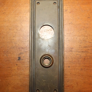 May include: A brass door plate with a round keyhole and two smaller holes. The plate has a decorative border and is rectangular in shape.