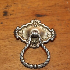May include: A silver-toned, ornate, decorative cabinet pull with a ring-shaped handle. The pull has a detailed, floral design and a smooth, polished finish.