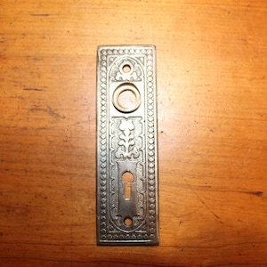 May include: A rectangular, antique-style door plate with ornate floral and geometric patterns. The metal plate has a keyhole, a circular knob opening, and two screw holes. The plate is silver-toned and sits on a wooden surface.