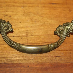 May include: Antique brass drawer pull with ornate, scroll-like detailing. The curved handle features decorative flourishes at each end, adding a vintage touch. The hardware is set against a wooden surface.