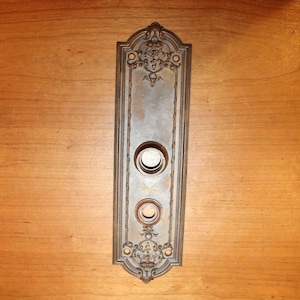 May include: A vintage-style door plate with ornate details and two round holes. The plate is made of metal and has a dark brown finish.