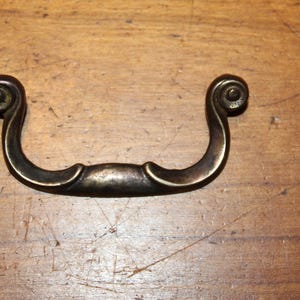 May include: Antique bronze-toned drawer pull with a curved handle and decorative scrollwork on each end. The hardware is set against a wooden surface, showcasing its vintage aesthetic. This piece is suitable for furniture restoration projects.