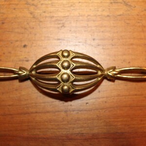 May include: A gold-toned metal drawer pull with a decorative, cage-like design and five round accents. The pull has a long, narrow shape with curved ends.