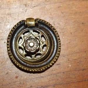 May include: A round, brass cabinet knob with a decorative, openwork design. The knob has a beaded edge and a ring pull.