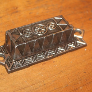 May include: A silver metal drawer pull with a geometric design. The pull has three circular decorative elements on the top.