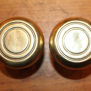 May include: Two round brass door knobs with concentric circles around the center. The knobs are polished and have a shiny finish.