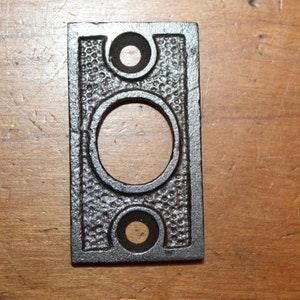 May include: A black metal door plate with a rectangular shape and two round holes. The plate has a textured surface.