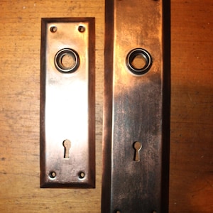 May include: Two antique copper door plates. The plates have a rectangular shape with rounded corners. Each plate has a keyhole and a circular opening for a door knob. The copper has a dark, aged patina.