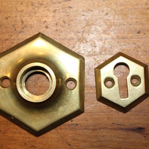 May include: Two antique brass door hardware pieces. One is a hexagonal plate with a central circular opening and two screw holes. The other is a smaller hexagonal escutcheon with a keyhole and two screw holes.