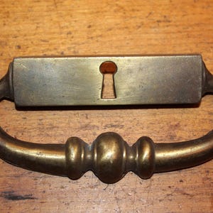 May include: A brass drawer pull with a keyhole in the center. The pull has a curved shape with a decorative knob in the center.
