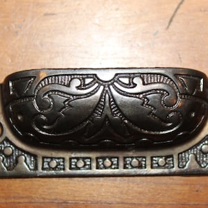 May include: A dark bronze-colored drawer pull with an ornate, symmetrical design. The handle features a central floral motif and decorative edges, with two holes for mounting. This hardware is suitable for cabinets and drawers.