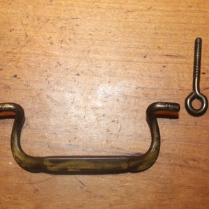 May include: A vintage brass drawer pull with two matching screws. The pull has a curved handle design and is likely from an antique cabinet or furniture piece. The screws have eyelets for attachment.