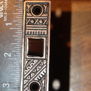 May include: A silver-colored rectangular metal object with intricate engraved patterns and two holes. The object is approximately 3 inches long and features a square inset and decorative designs.