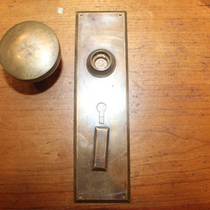 May include: A brass door knob and backplate with a keyhole. The knob is round and has a smooth surface. The backplate is rectangular and has a raised edge.