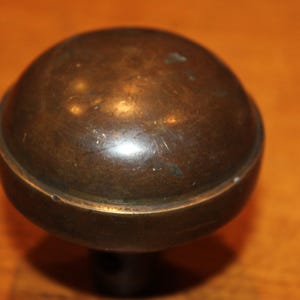 May include: A close-up of a bronze-colored spherical cabinet knob. The knob has a rounded top and a circular base, with a stem extending downwards. The surface shows signs of wear and age, with a slightly tarnished appearance.