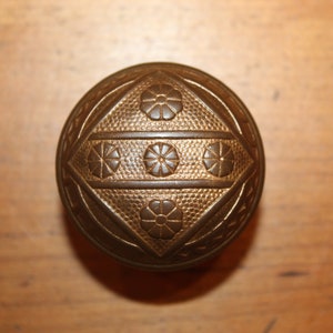 May include: A round, brass door knob with a geometric design and floral accents. The knob has a textured surface and a raised, diamond-shaped pattern.