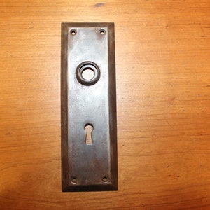 May include: A vintage, rectangular, metal door plate with a keyhole and a round knob hole. The plate is a dark brown color and has a worn, aged appearance.