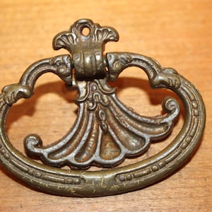 May include: An ornate brass drawer pull with a decorative, fan-shaped design in the center. The pull has a rounded oval shape with intricate details.