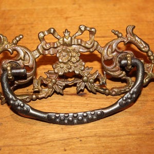 May include: Ornate antique drawer pull with a decorative floral and scroll design. The pull features a dark, curved handle with a series of small, raised dots. The metal has a gold and bronze finish, adding to its vintage appeal.