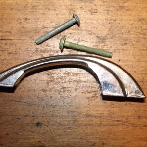 May include: A chrome-finished cabinet pull with a curved design, accompanied by two screws. The hardware is set against a wooden surface, suggesting a vintage or antique aesthetic. Ideal for furniture restoration.