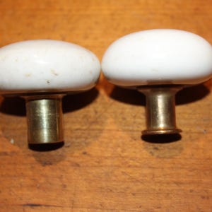 May include: Two vintage white ceramic cabinet knobs with brass stems. The knobs have an oval shape and are slightly aged, showing subtle imperfections. The brass stems add a classic touch to these decorative hardware pieces.