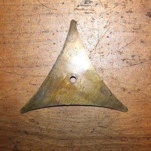 May include: A brass triangle-shaped object with a hole in the center. The object has a dull, aged appearance and is set against a wooden background. The triangle has rounded corners and a pointed top.