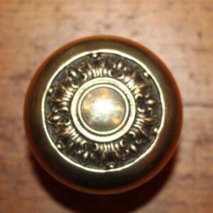 May include: A round, antique brass door knob with an ornate floral design around the center. The knob has a polished finish and is set against a wooden background.