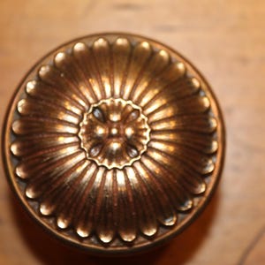 May include: Close-up of a vintage brass doorknob with a detailed, floral-like design. The knob has a circular shape with a central rosette and radiating petal-like edges. The brass has a warm, aged patina.