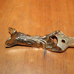 May include: A gold metal furniture leg with a decorative floral design. The leg has two holes for mounting.