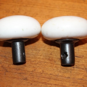 May include: Two white ceramic door knobs with black metal stems. The knobs are round and have a smooth finish.